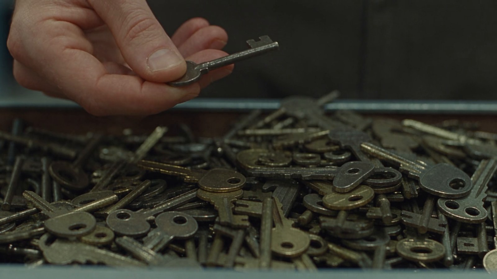 A hand digs through a bargain bin of loose old keys, picking one up and dropping it back.